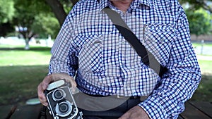 Aged man is sitting on bench in park at summer day, taking retro photo camera