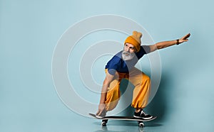 Aged man in t-shirt, orange pants, hat, gumshoes. Riding black skateboard, looking at it, posing on blue background