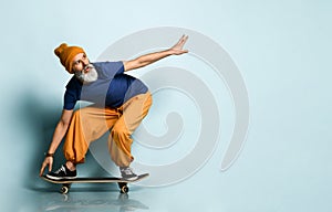 Aged man in t-shirt, orange pants, hat, gumshoes. Riding black skateboard, looking at it, posing on blue background