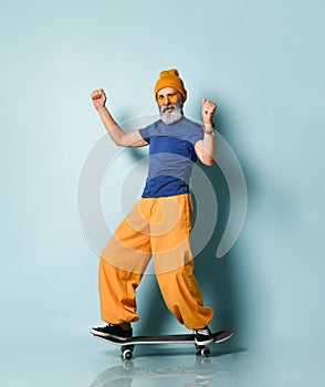 Aged man in t-shirt, sunglasses, orange pants, hat, gumshoes. Riding black skateboard, showing fists, posing on blue background