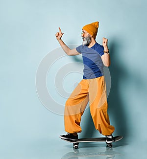 Aged man in t-shirt, sunglasses, orange pants, hat, gumshoes. Riding black skateboard, showing fists, posing on blue background