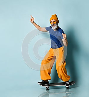 Aged man in t-shirt, sunglasses, orange pants, hat, gumshoes. Riding black skateboard, showing fists, posing on blue background