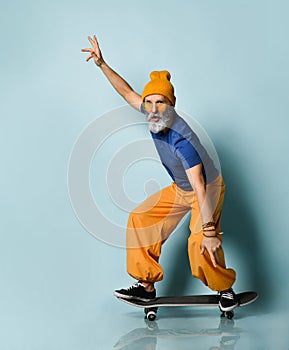 Aged man in t-shirt, sunglasses, orange pants, hat, gumshoes. Riding black skateboard, showing fists, posing on blue background