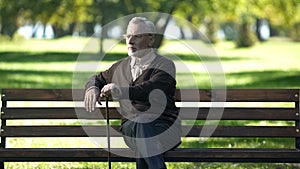 Aged man with walking stick resting on bench in park, enjoying spring nature