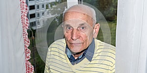 Aged man in yellow pullover stands at white curtains