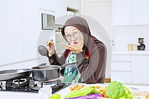 Aged Muslim woman tasting soup from pot at home