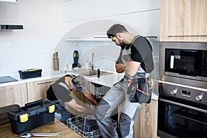 Aged repairman in uniform fixing dishwasher in the kitchen, while his young colleague standing near him and watching