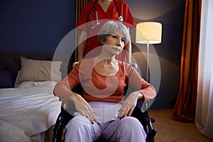 Aged senior woman in wheelchair having professional help at nursing home