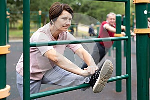 Aged woman doing stretching on sports bars in open-air sports area