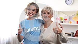 Aged woman and female volunteer showing thumbs up and hugging, retiree help