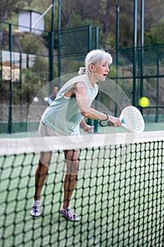 Aged woman hitting ball with backhand during paddleball match on outdoor court