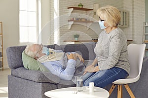 Aged woman in medical protective mask taking care of elderly husband feeling fever