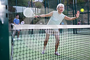 Aged woman playing friendly paddleball match on outdoor court