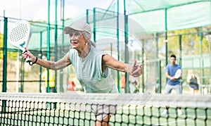 Aged woman playing friendly paddleball match on outdoor court