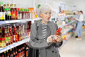 Aged woman reading labels on bottles with sauces in supermarket