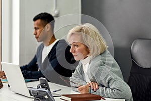 Aged woman, senior intern looking focused at the screen while using laptop, sitting at desk, working in modern office