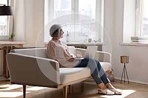 Aged woman sit on sofa with laptop staring out window