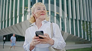 Aged woman smartphone looking around at urban street waiting meeting close up.