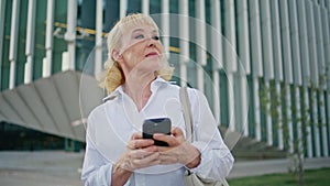 Aged woman smartphone looking around at urban street waiting meeting close up.