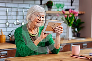 Aged woman tenderly palming photo frame while sitting at table