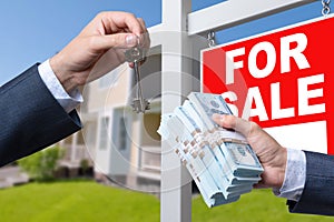 Agent Handing Over Keys as Buyer is Handing Over Cash for House with Home and For Sale Real Estate Sign Behind.