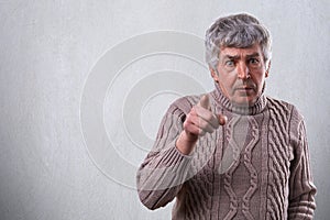 An aggressive adult man with gray hair and wide open eyes pointing at you with finger. Emotional senior man wearing sweater standi