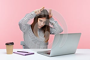 Aggressive business woman holding fingers above head doing bull horn gesture and looking at laptop screen with anger, threatening