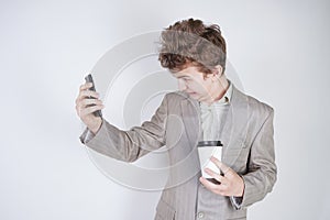 Aggressive caucasian teenager screams into the smart phone and holds a paper cup of coffee in his hand. student on edge on a white