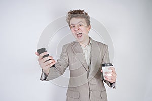 Aggressive caucasian teenager screams into the smart phone and holds a paper cup of coffee in his hand. student on edge on a white