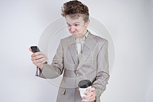 Aggressive caucasian teenager screams into the smart phone and holds a paper cup of coffee in his hand. student on edge on a white