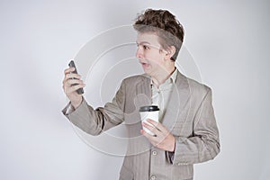 Aggressive caucasian teenager screams into the smart phone and holds a paper cup of coffee in his hand. student on edge on a white