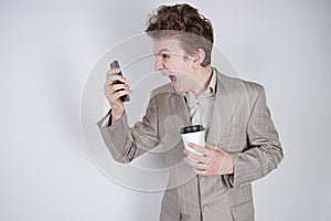 Aggressive caucasian teenager screams into the smart phone and holds a paper cup of coffee in his hand. student on edge on a white