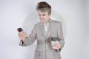 Aggressive caucasian teenager screams into the smart phone and holds a paper cup of coffee in his hand. student on edge on a white