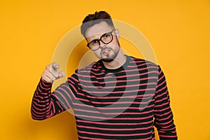 Aggressive man in striped sweatshirt and eyeglasses pointing at something on yellow background