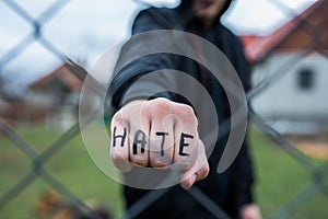 Aggressive teenage boy showing hes fist behind wired fence at the correctional institute, the word hate is written on hes hand