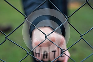 Aggressive  teenage boy showing hes fist behind wired fence at the correctional institute, the word hate is written on hes hand