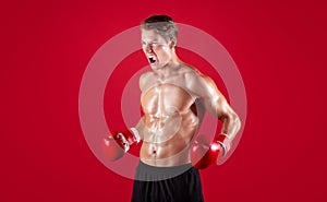 Aggressive young sportsman in boxing gloves screaming in anger on red studio background