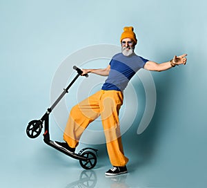 Aging man in t-shirt, sunglasses, orange pants, hat, gumshoes. Riding black scooter, smiling, posing on blue background