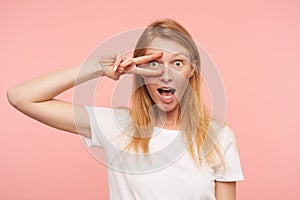 Agitated young attractive redhead lady with natural makeup keeping victory gesture near her face while looking excitedly at camera