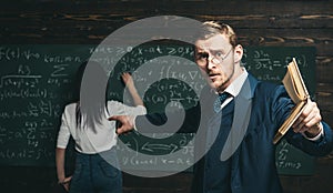 Agitated young professor with stylish mustache and beard pointing book at students in classroom while showing formula