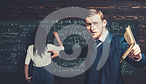 Agitated young professor with stylish mustache and beard pointing book at students in classroom while showing formula