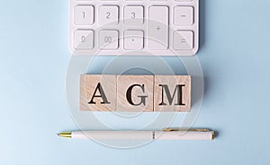 AGM word on wooden block with pen and calculator on blue background