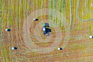 An agricultural tractor collects mowed grass for agricultural use and wraps hay bales in a plastic field, aerial top view