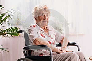 Ailing woman dressed in a blouse with flower pattern is sitting in a wheelchair in her home