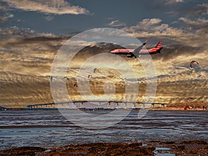 Aircraft flits over a bridge and many surf kites at dusk photo