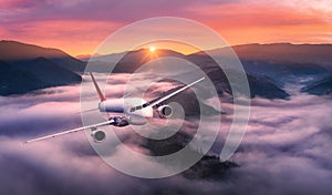 Airplane is flying above mountain peak in pink low clouds