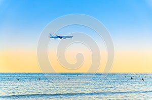 Airplane flying low above sea and people tourists swimming in water, clear blue orange sky at sunset, plane preparing to land