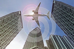 Airplane flying over high rise buildings with bright sunlight in background.