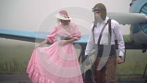 An airplane pilot in a vintage helmet and a girl in a pink dress and a white hat stand near the plane in a fog, slow