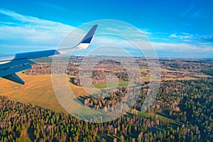 Airplane window view. View from the window on the spring forest, meadows, blue sky. Airplane`s wing flying above the colorful land